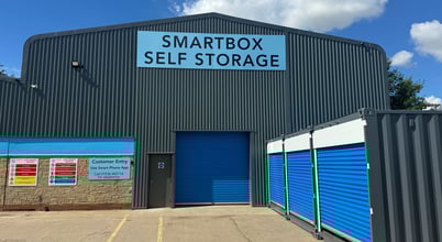 The image shows a warehouse building labeled "SMARTBOX SELF STORAGE" with a large blue roll-up door. There are two shipping containers with blue doors adjacent to the building. Various signs, including safety instructions and customer entry instructions, are displayed on the building's side. The ground is paved, and the sky is clear with some clouds.