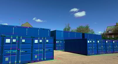 A sunny outdoor storage facility with several blue shipping containers arranged in rows. The containers are stacked and have numbers and labels on them. In the background, a clear blue sky with a few clouds and some trees and rooftops are visible.