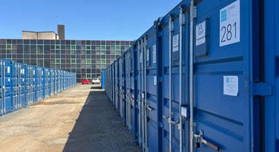 A row of blue shipping containers aligned on both sides of a gravel pathway with a clear blue sky above. In the background, there's a building with multicolored panels on its facade.