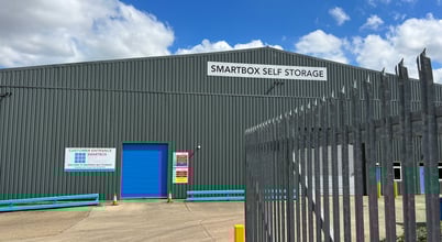 A large warehouse building with a sign reading 'Smartbox Self Storage' on its facade. The structure has a corrugated metal exterior with a bright blue roll-up door. There is a fenced area in the foreground and a clear blue sky with some clouds in the background.