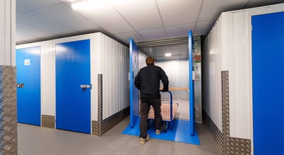 A person pushing a trolley into a storage unit facility, characterized by blue doors and metal walls. The area is well-lit, and a fire exit sign is visible on the ceiling.