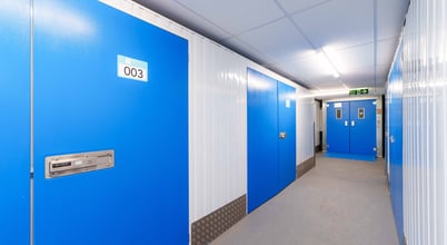 A corridor in a storage facility with bright blue doors on either side and a gray floor. The door closest to the foreground is labeled with the number '003'. Overhead, fluorescent lights illuminate the space, and there is an exit sign at the end of the corridor above double doors.