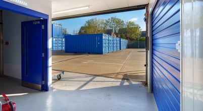 View from inside a storage facility with a partially open door leading to an outdoor area filled with several blue shipping containers. The ground is paved and a clear sky is visible above.