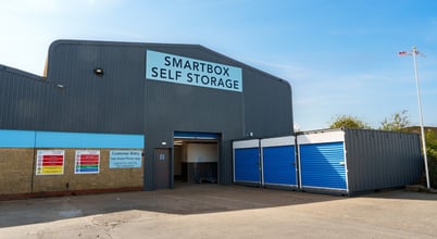 A large grey industrial building with a sign reading 'Smartbox Self Storage'. The building has blue accents and an open entrance. Nearby are blue storage units. Various informational signs are displayed on the wall. The sky is clear and blue.