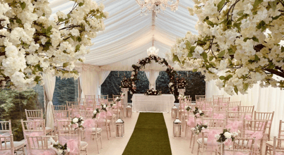 A beautifully decorated wedding venue inside a white tent. The aisle is adorned with green artificial grass and lined with chairs decorated with pink sashes and floral arrangements. Large white floral displays and chandeliers hang from the ceiling, with a floral arch visible at the end of the aisle in front of a table.