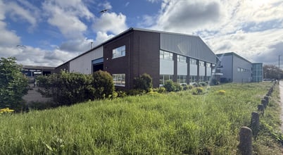 A large industrial-style building on Howard Road, Eaton Socon, St Neots with a brick and metal facade, surrounded by lush green grass and shrubs. The sky above is partly cloudy with sunlight streaming through, creating a bright and crisp atmosphere. Birds can be seen flying in the sky, and there are bollards lining the grassy area.