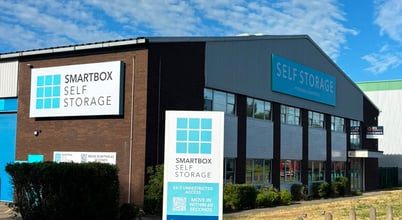 A self-storage facility with a sign reading 'Smartbox Self Storage'. The building has a blue and white color scheme with large windows. There is a billboard in front displaying similar branding and contact information for the storage services. The sky is clear with scattered clouds.