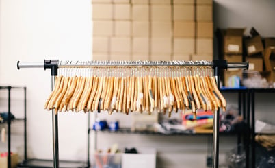 A clothing rack with wooden hangers arranged closely together. In the background, there are shelves and stacked boxes, suggesting a storage or warehouse setting.