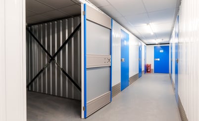 A well-lit hallway in a self-storage facility, featuring several blue storage unit doors. One unit door is open, revealing an empty storage space with metal walls and diagonal support beams. The floor and walls are clean, and a fire extinguisher is visible at the end of the hallway.