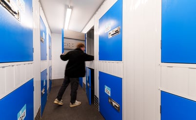 A person in a black jacket standing in a hallway with blue and white storage lockers. The person is seen from the back, holding a door open to one of the lockers. The hallway is narrow, with fluorescent lighting above and metal floors below.