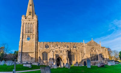A historic stone church with a tall spire and clock on the tower, surrounded by a graveyard with tombstones and green grass, set against a clear blue sky.