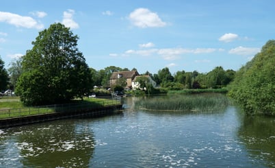 A tranquil river scene in Eaton Socon with calm water reflecting the blue sky and fluffy clouds. Surrounding the river are lush green trees and grassy banks. A small building with a sloped roof is visible in the background, partially obscured by trees.