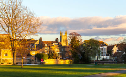A scenic view of St Neots at sunset, featuring a green field, leafless trees, and historic stone buildings bathed in warm golden light. In the background, a tall church tower rises above the rooftops. The sky is partly cloudy with soft pink and blue tones, adding to the tranquil atmosphere.