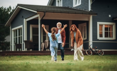 A young girl runs towards an elderly couple standing on the grass in front of a blue house. The girl wears overalls and has her arms outstretched, while the elderly man and woman are smiling, with the man extending his arms to greet her. The house has large windows and a bicycle is visible in the background.