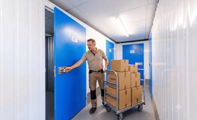 A man pushing a cart loaded with cardboard boxes labeled 'Smartbox' in a storage facility. He is opening a blue door labeled '007' to a storage unit. The corridor is lined with white walls and fluorescent lighting.