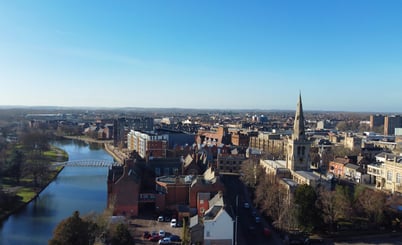 Aerial view of Bedford with a river running through it. To the right, a prominent church with a tall spire stands among other historic buildings. Modern structures and residential areas are visible in the background. A bridge crosses the river, surrounded by green spaces and trees. The sky is clear and blue.