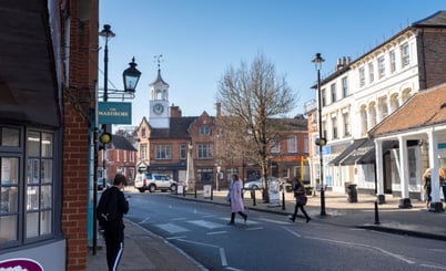 A street scene in Ampthill, UK with a blend of traditional and modern architecture. There is a brick building with a sign reading 'The Wardrobe' on the left, and more buildings on the right side of the road. A clock tower is visible in the background, and several people are walking along the street under clear blue skies.