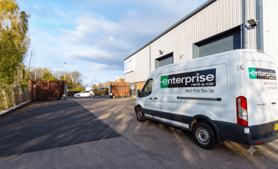 A white van labeled 'Enterprise Rent-A-Car' parked outside a commercial building with large garage doors. The van has a slogan 'We'll Pick You Up' written on it. The parking area is surrounded by fencing and there are some trees and a few cars in the distance. The sky is partly cloudy with some blue patches visible.