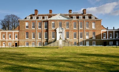A large, historic brick mansion with numerous white-framed windows and chimneys on the roof, set against a blue sky. The building features a central entrance with a pediment above and is flanked by side wings. In front of the mansion is a well-maintained lawn and a decorative statue on a plinth.