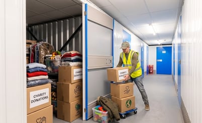 A person wearing a neon yellow safety vest and cap stacks boxes labeled "Charity Donations" in a storage facility hallway. The storage unit is open, revealing clothing and toys inside. The hallway has white walls, blue doors, and overhead lighting.