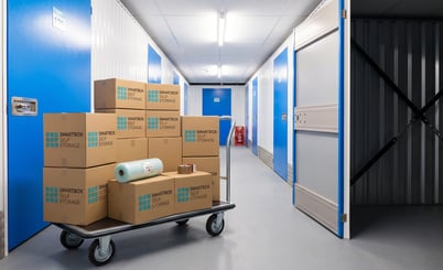 A self-storage facility corridor with blue and white doors. A cart with several cardboard boxes labeled 'Smartbox Self Storage' is placed in the hallway. Some packing materials are on top of the boxes. One of the storage unit doors is open, showing an empty metal interior. Overhead fluorescent lights illuminate the scene.