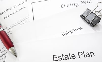 A close-up of several documents related to estate planning. Visible text includes 'Estate Plan,' 'Living Trust,' 'Living Will,' and 'Healthcare Power of Attorney Advance Directive.' There is a red pen resting on one document and a black binder clip on another.