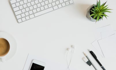 A tidy workspace featuring a white keyboard, a potted succulent plant, a cup of coffee, a smartphone, earphones, a pen, a pencil, an eraser, a USB flash drive, and scattered sheets of white paper, all on a white surface.