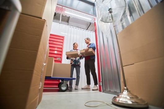 Two people are standing in a storage unit filled with cardboard boxes. One person is holding a box, and they appear to be smiling at each other. A stack of boxes and a dolly are visible in the foreground, and a floor lamp is on the right side.