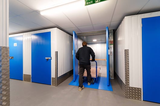 A person pushing a trolley into an elevator in a storage facility with blue and white doors and units. The floor is covered with a textured metal plate, and an exit sign is visible on the ceiling.