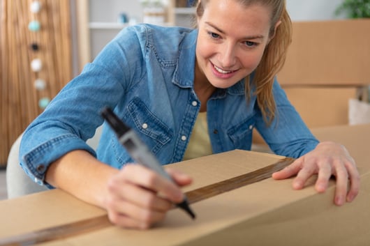 A young woman wearing a denim shirt is smiling and writing on a cardboard box with a black marker. She is indoors, with more boxes visible in the background, suggesting a moving or packing scenario.