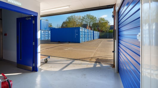 An open storage unit with a blue door looking out onto an outdoor area at Desborough filled with multiple blue storage containers stacked and aligned. The sky is clear, and there are trees and a fence visible in the background.