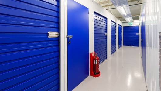 A corridor in a storage facility with several blue metal storage unit doors. The doors have padlocks and the walls are light-colored. A red fire extinguisher is mounted on the wall at the entrance. Overhead fluorescent lighting illuminates the hallway.