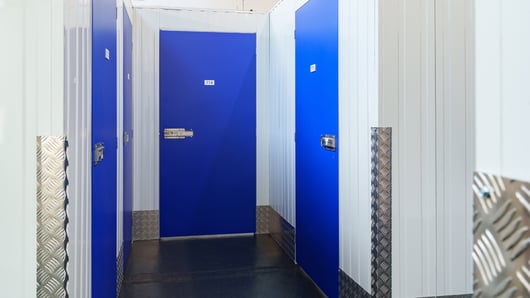 A corridor with several blue doors leading to storage units, each door is numbered and secured with a latch. The walls are white with metal ridges, and the floor is dark, with metal protector panels on the lower parts of the walls.