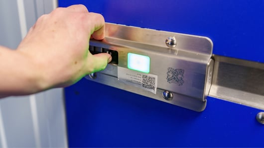 A person inserting a key card into a metallic card reader attached to a blue door. The card reader displays a green light, indicating successful access. The setup includes a QR code and a logo engraved on the metallic surface.