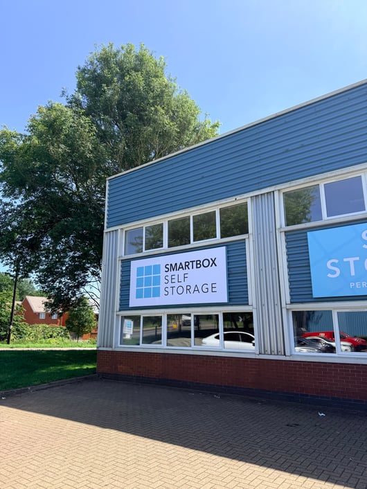 A building exterior with a sign that reads 'SMARTBOX SELF STORAGE' on a blue background. The building has blue corrugated metal siding with large windows and is situated next to a tree. A car is partially visible through the windows.