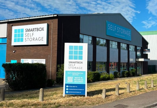 A commercial building with signs that read 'Smartbox Self Storage St Neots’ and 'Self Storage Personal &amp; Business.' The building is brown and has several large windows. There are shrubs and a pathway leading to the entrance. A large exterior sign provides contact information and highlights 24/7 access and quick move-in availability.