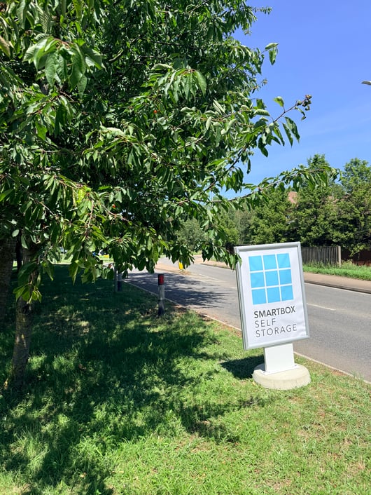 A sign for Smartbox Self Storage is placed on a grassy area beside a road. The sign features a blue grid pattern above the text. A lush green tree with abundant leaves and small red fruit is adjacent to the sign, partially casting a shadow on the lawn. The road is lined with more greenery and trees, and the day is sunny with a clear blue sky.