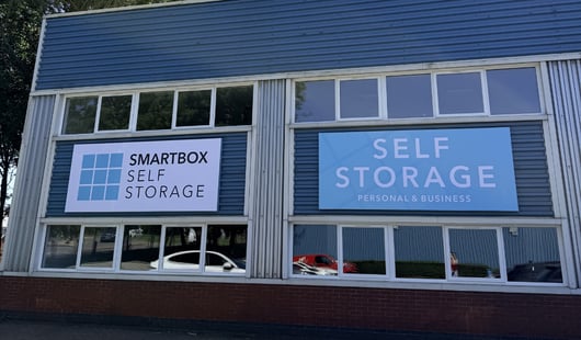 A Smartbox Self Storage Ampthill with two signs, one reads 'SMARTBOX SELF STORAGE' with a grid logo on the left, and the other reads 'SELF STORAGE PERSONAL &amp; BUSINESS'. The building has blue metal siding and large windows.