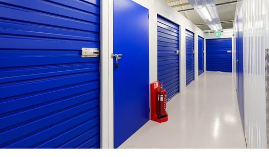 A well-lit storage facility hallway with blue roll-up doors and white walls. A fire extinguisher is positioned on the floor next to one of the doors. The ceiling has fluorescent lighting, and an exit sign is visible in the distance.