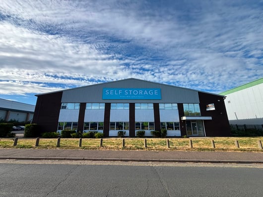 A large industrial building with a sign reading 'SELF STORAGE Personal & Business' on the front. The structure is dark-colored with a row of windows and a small entrance. It is surrounded by a grassy area and situated near a paved road. The sky above is mostly cloudy with patches of blue.