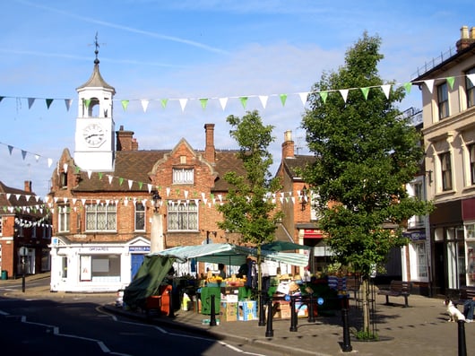 A quaint town square in Ampthill with a traditional brick building and a clock tower. The building has an old-style architecture with a gable roof and decorative windows. In the foreground, there is a market stall covered with a green and white canopy selling fruits and vegetables. Festive bunting is strung across the square, and there are a few trees and benches in the area. The sky is clear and blue, suggesting a sunny day.